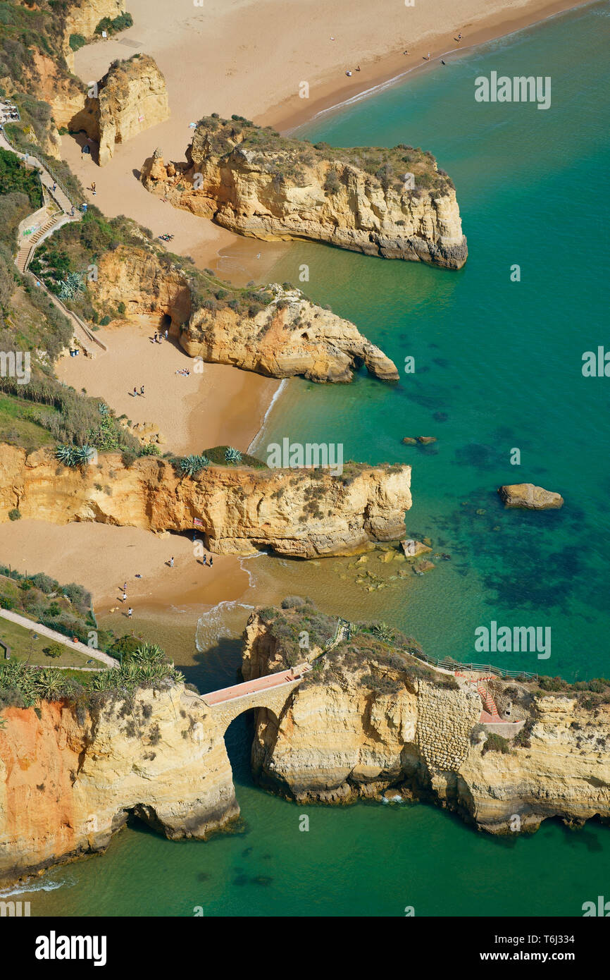 VISTA AEREA. Serie di scogliere parallele sul mare a Praia Dos Estudiantes (spiaggia degli studenti), ponte romano in primo piano. Lagos, Algarve, Portogallo. Foto Stock