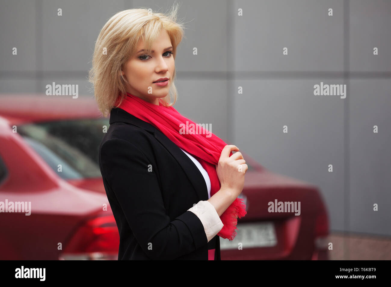 Moda giovane donna d'affari accanto alla sua auto elegante modello femminile indossando abito nero giacca e sciarpa rossa all'aperto Foto Stock