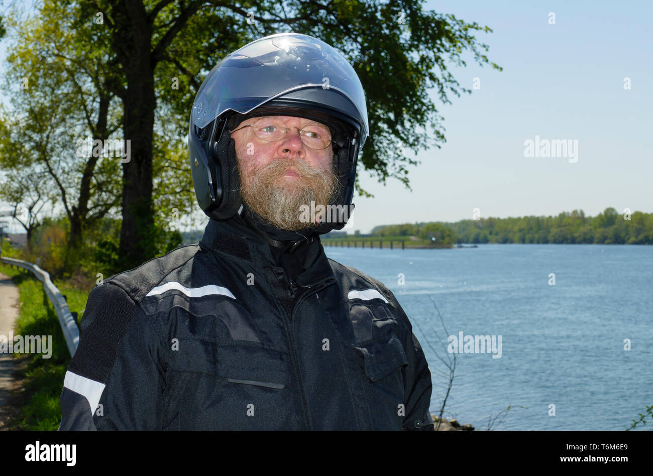 Mann mit langem Bart, Brille und Motorradhelm, steht am Ufer des Rhein Foto Stock
