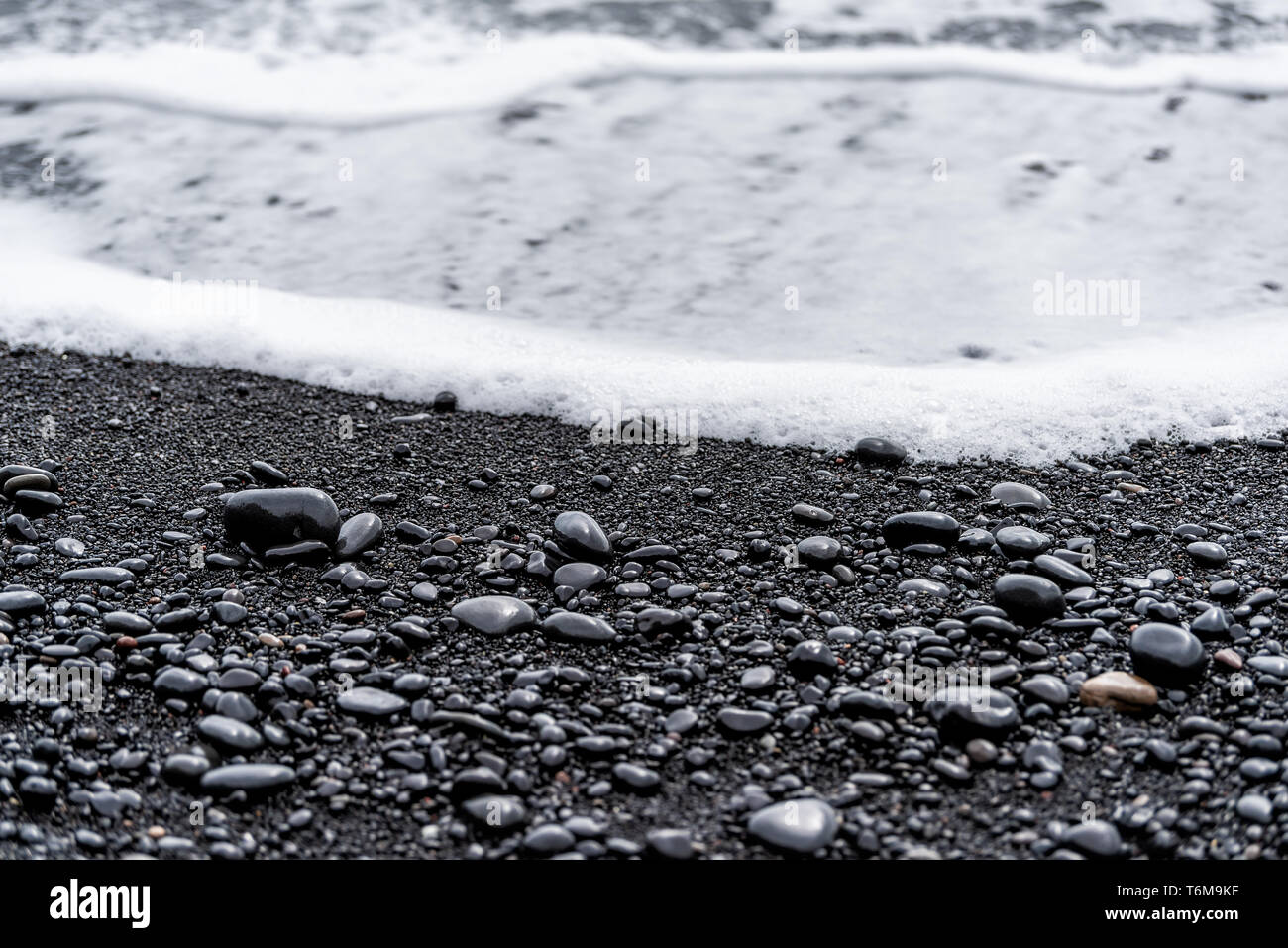 Nero pietre vulcaniche sulla spiaggia di sabbia in Reynisfjara, Islanda con acqua onde che si infrangono sulla riva a lucido rocce umide in Vik Foto Stock