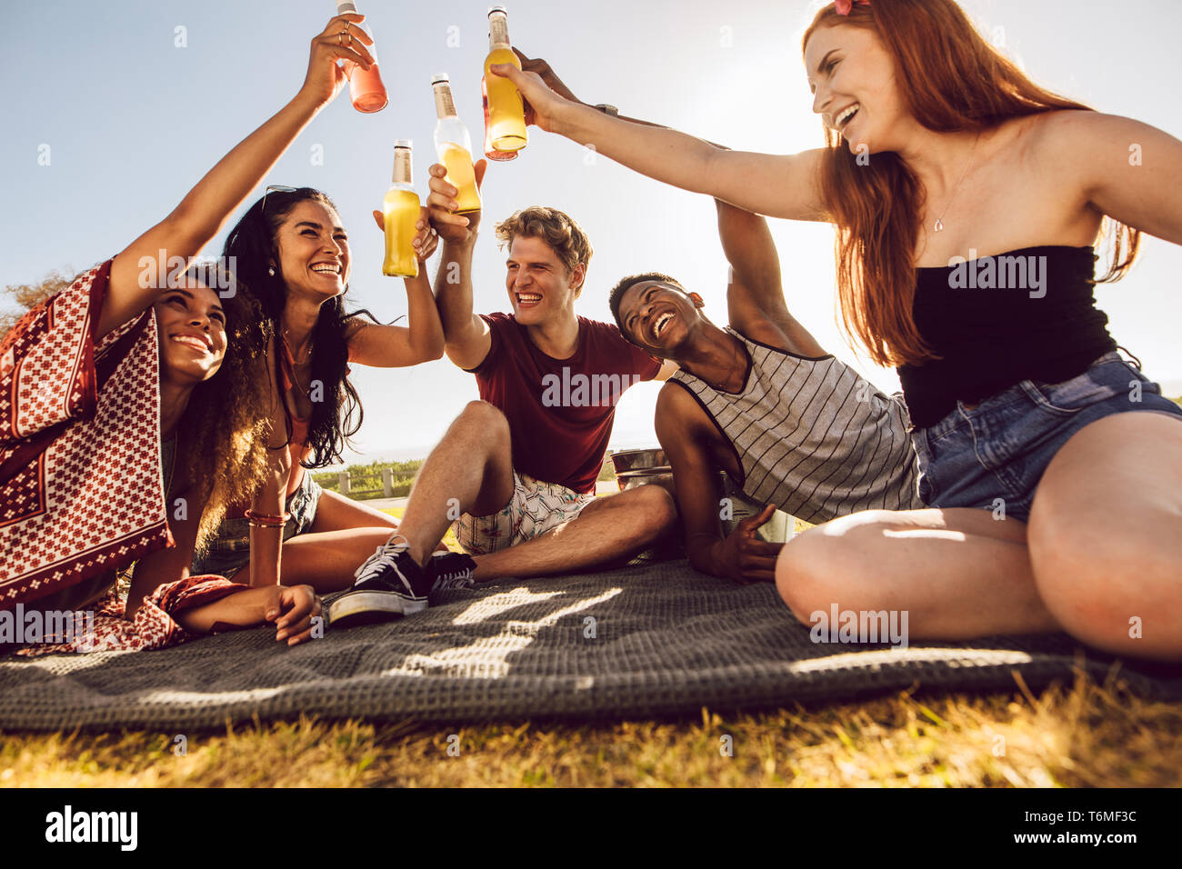 Multi-etnico amici seduti all'aperto su un plaid e tostatura di birra. Giovani uomini e donne aventi una festa all'aperto su un giorno d'estate. Foto Stock