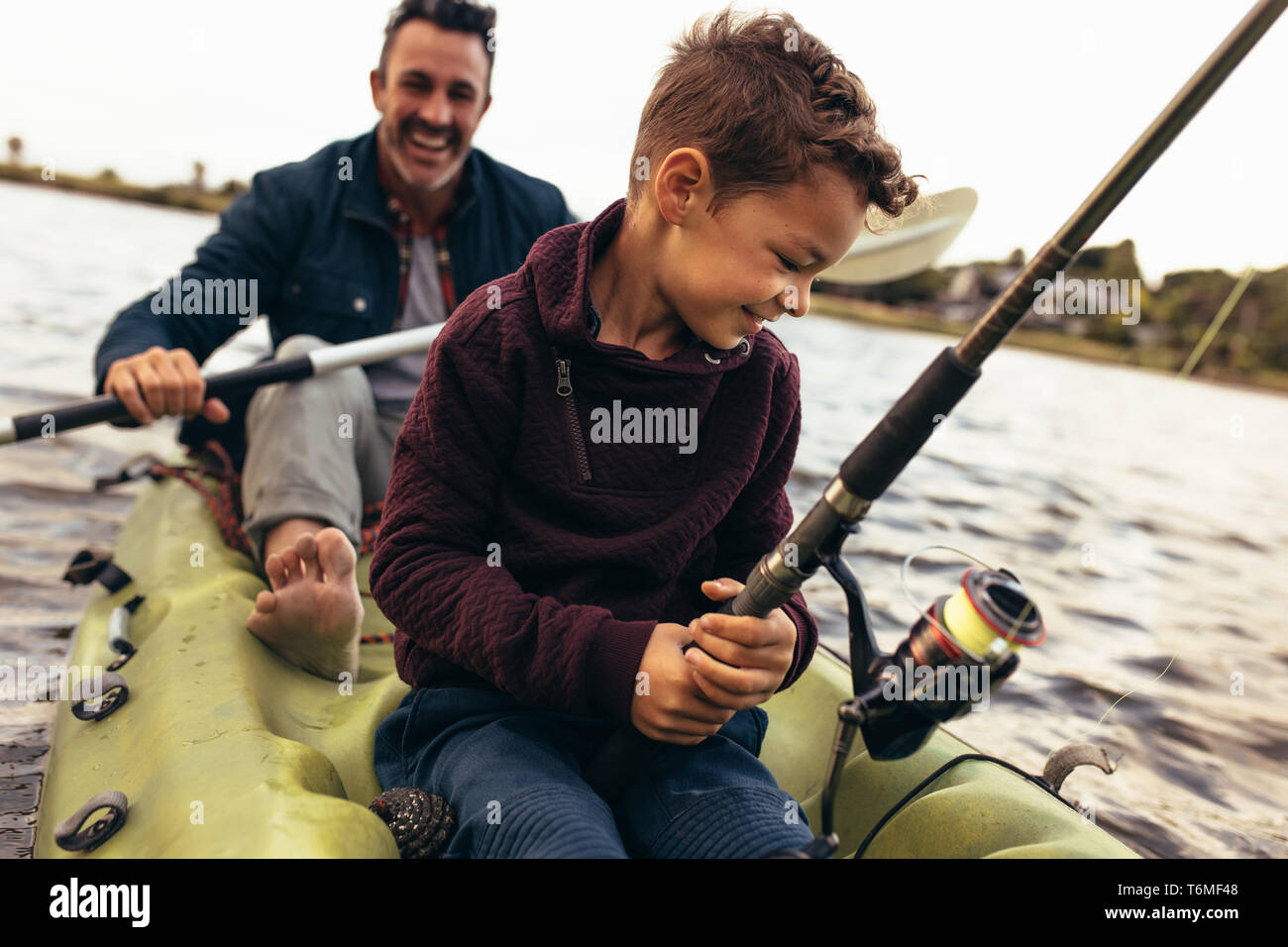 Close up di un bambino seduto in un kayak per la cattura di pesce in possesso di una canna da pesca. Uomo felice canottaggio una piccola barca in un lago mentre il suo ragazzo cerca di catturare pesci us Foto Stock
