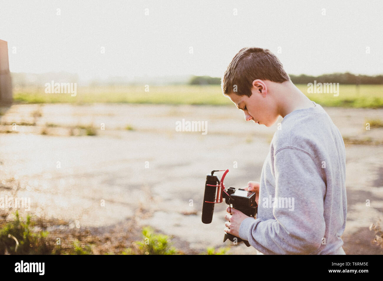 Giovane maschio videografo studiando e utilizzando la fotocamera mirrorless con microfono accessorio per la creazione di un video. Tramonto con candelette mette in evidenza il contrasto. Foto Stock