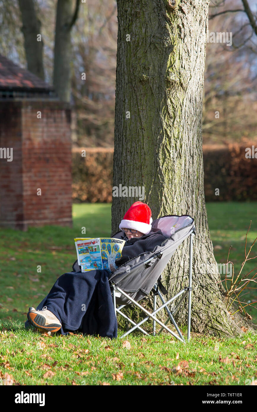 Ragazzo isolato nel parco da albero indossando cappello di Santa, seduto in sedia da campeggio, avvolto in coperta di vello, sorridente & libro comico di lettura in inverno sole. Foto Stock