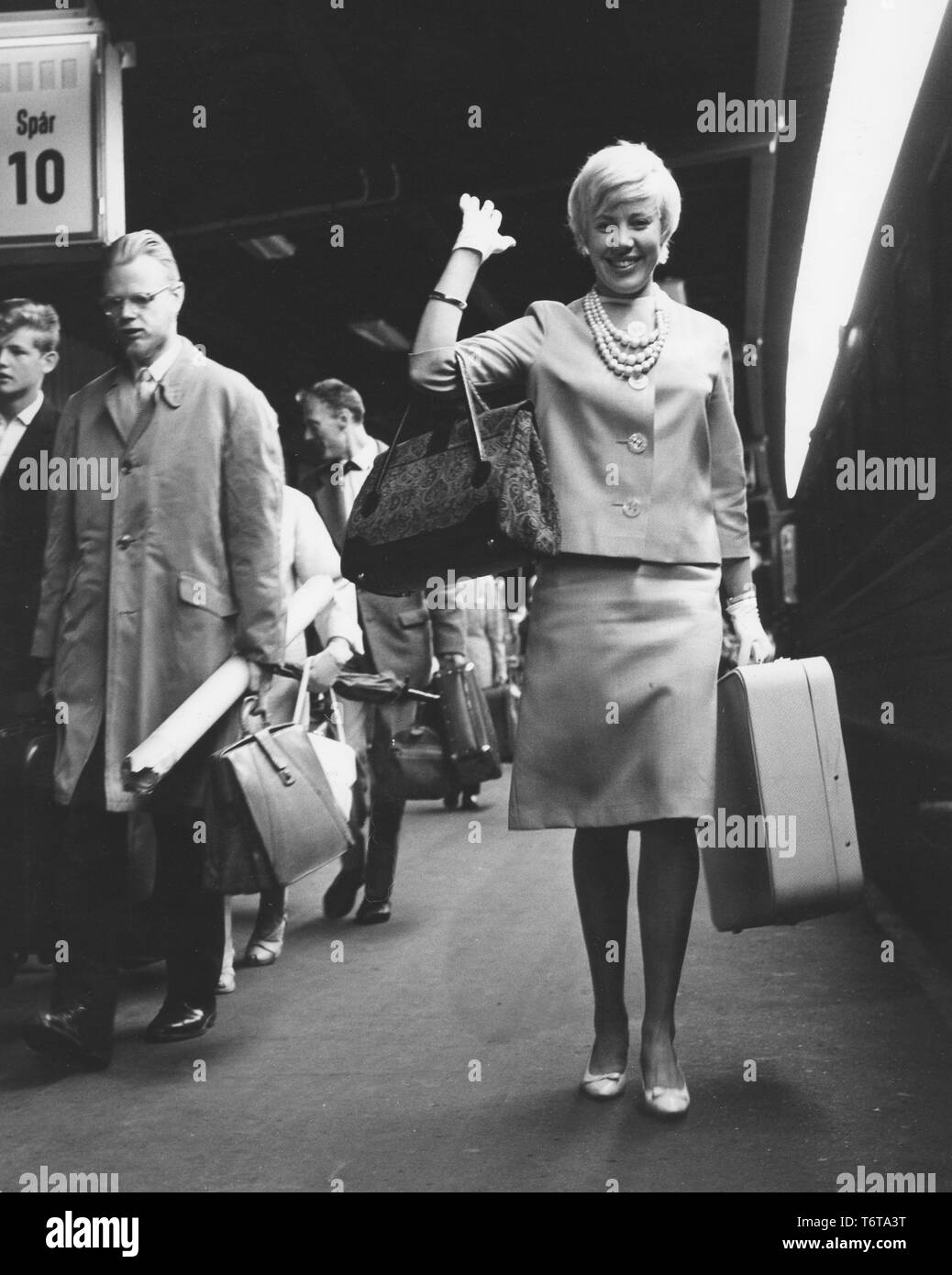 Viaggiare in treno negli anni sessanta. Una giovane donna bionda arriva alla stazione dei treni di onde e felicemente. Ella è ornate graziosamente in un tipico a due pezzi gonna e giacca. Ella è anche indossando una collana di adattamento e i guanti bianchi. La Svezia 1961 Foto Stock