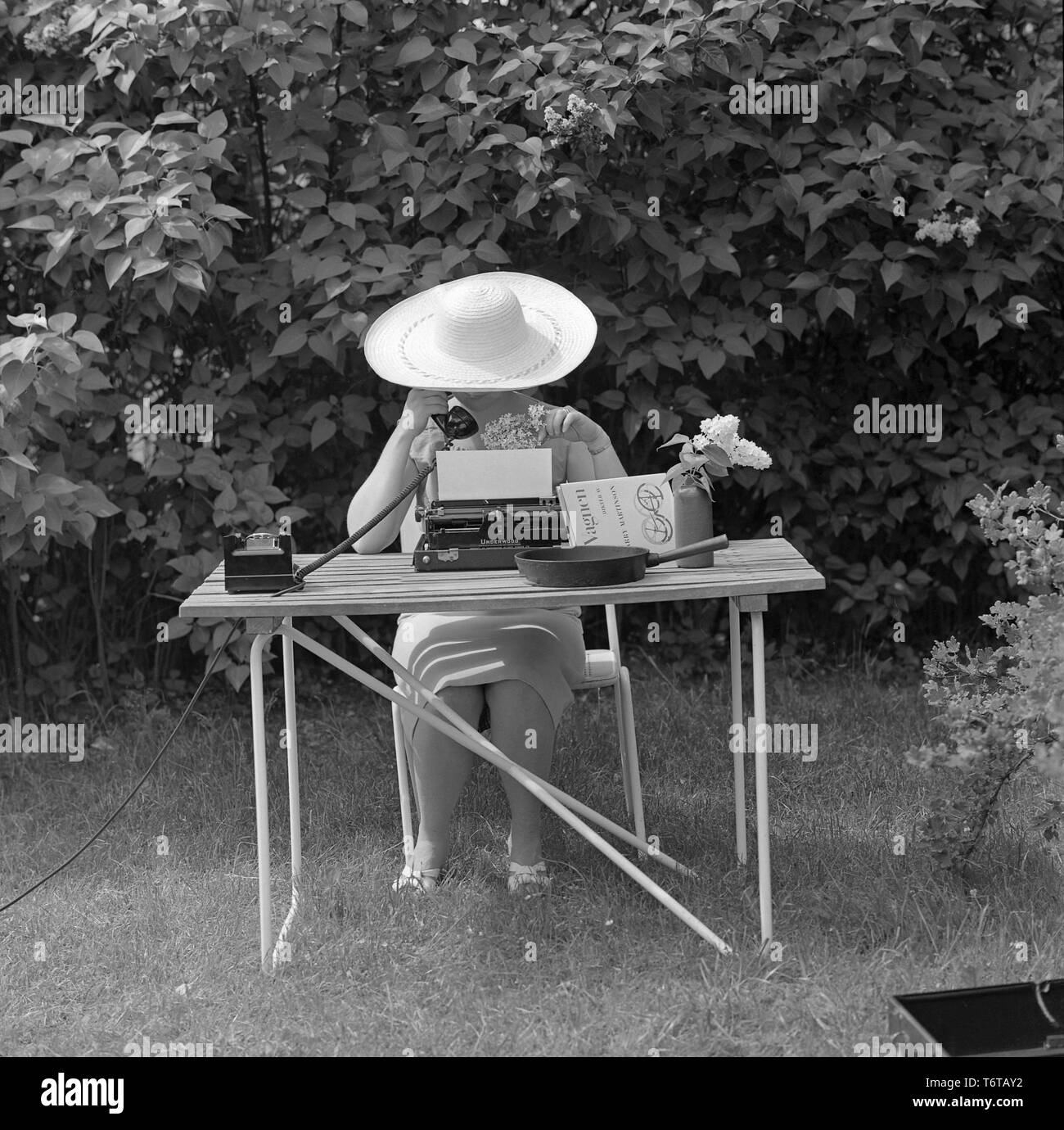 Estate negli anni '1960 Una signora con un cappello è seduta in giardino a scrivere su una macchina da scrivere. Allo stesso tempo sta parlando al telefono. Una supposizione è che stia cercando di fare un po' di lavoro durante la sua vacanza. Svezia 1961 Foto Stock