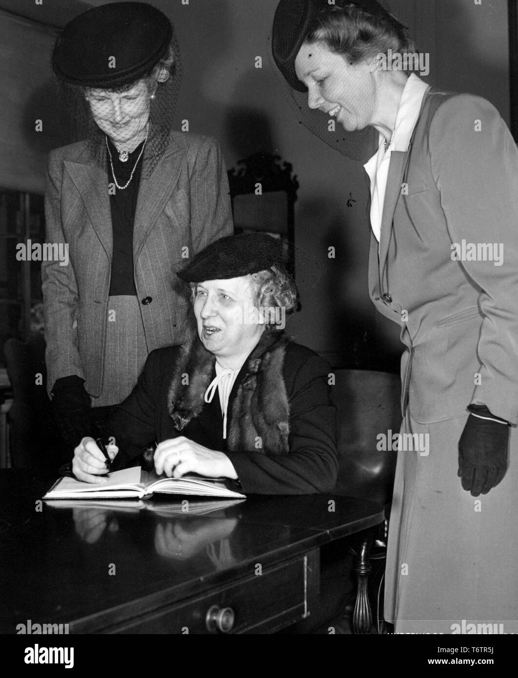 La First Lady Bess Truman, seduti ad una scrivania, cerca di scrivere in un libro mentre due donne sorridenti, indossa una scatola di pillole reticolare cappelli, guardare oltre la sua spalla, 1946. Immagine cortesia archivi nazionali. () Foto Stock