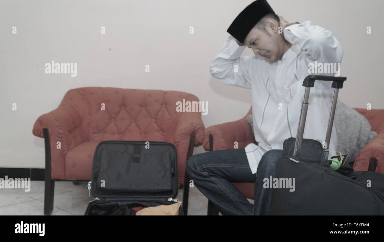 Musulmani uomo asiatico con songkok o cappuccio di testa seduto sul divano rosso ha sottolineato preparare per viaggiare con roba su nero valigia bagagli - immagine Foto Stock