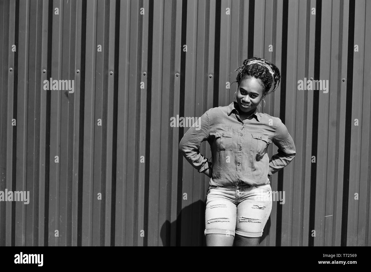 Donna africana con i timori dei capelli, in jeans corti poste contro verde parete in acciaio. Foto Stock