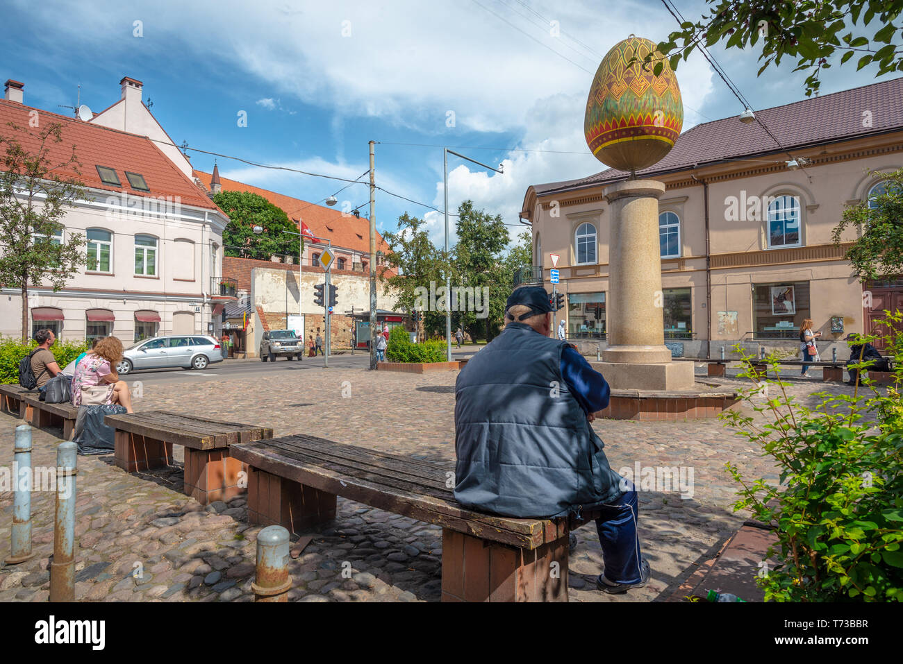 Un uomo e altre due persone sono seduti in una piccola piazza in cui sorge una scultura di un uovo decorato a Vilnius, Lituania Foto Stock