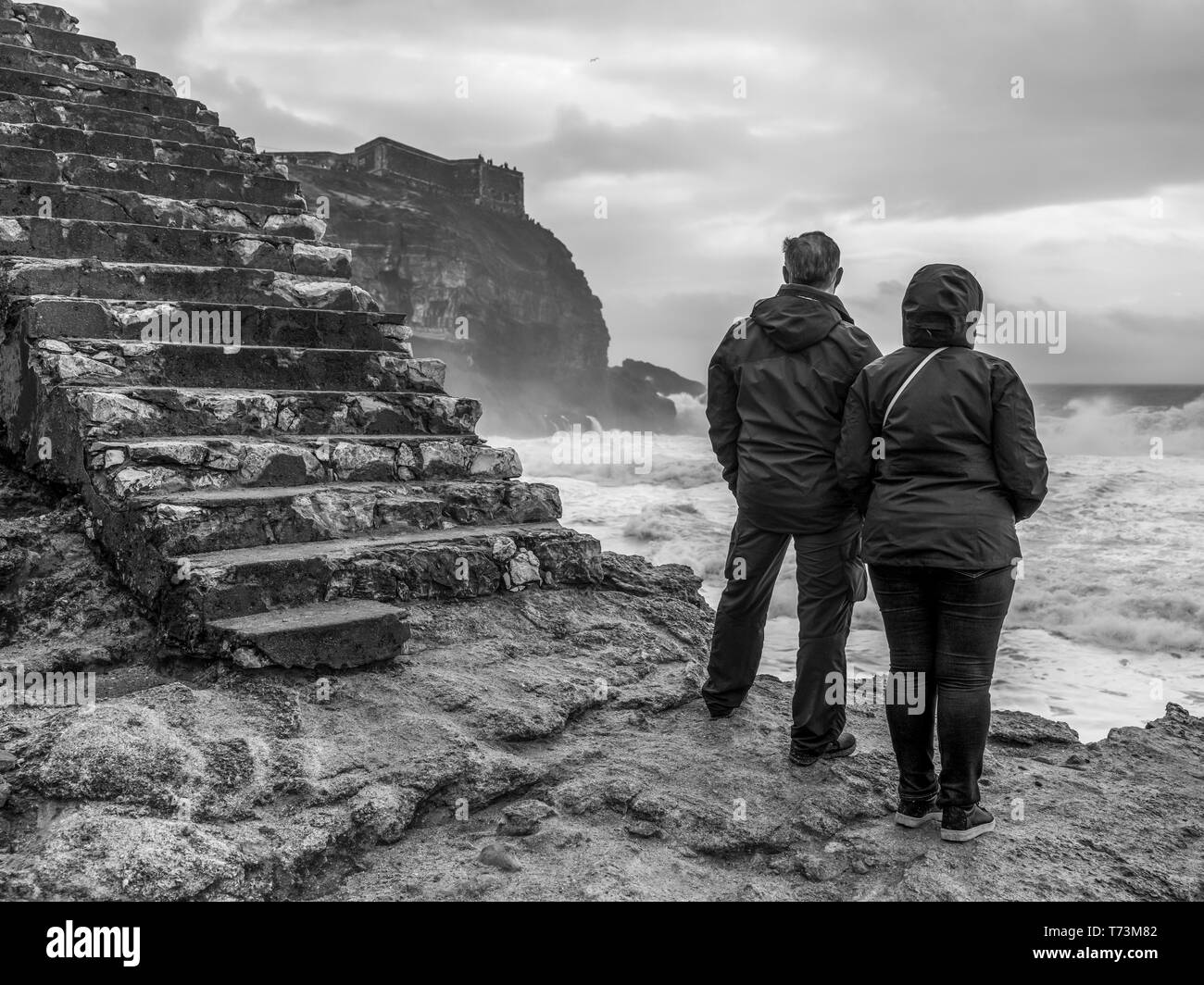 Un giovane si erge sul bordo dell'acqua che guarda al mare mosso da Nazare Beach, una popolare località balneare; Nazare, Distretto di Leiria, Portogallo Foto Stock