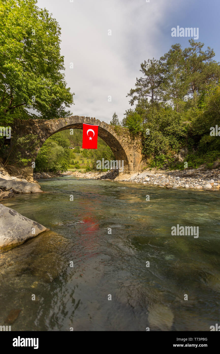 Il vecchio ponte di pietra sul fiume di montagna con acqua cristallina e la bandiera della Turchia appesi da quel ponte. Composizione verticale. Foto Stock