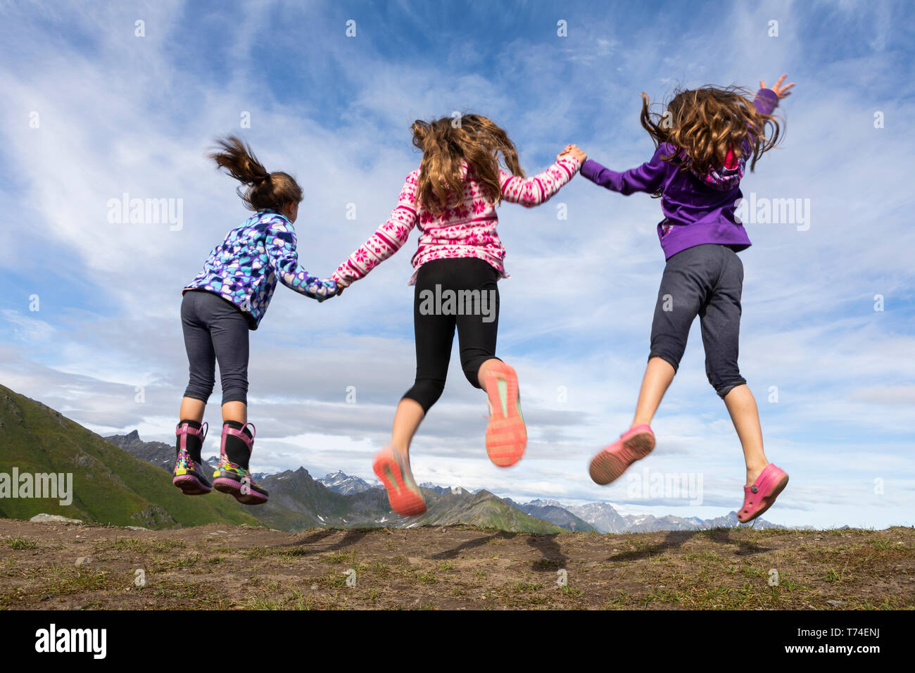 Tre giovani sorelle di salto e tenendo le mani nel Talkeetna montagne, Hatcher's Pass; Alaska, Stati Uniti d'America Foto Stock
