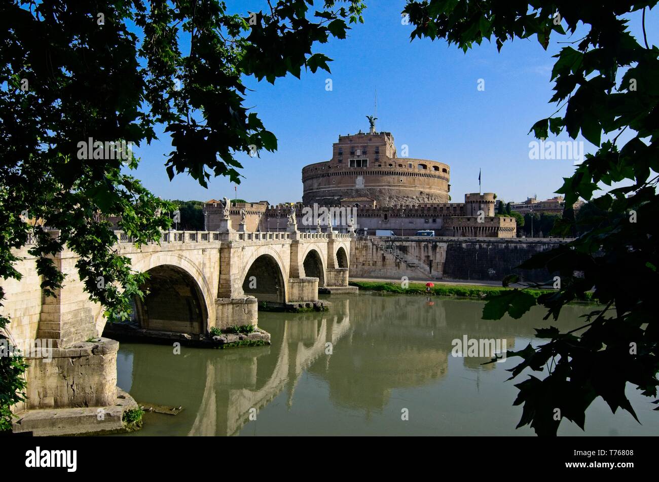 Vista attraverso gli alberi di Ponte Sant'Angelo e Castel Sant'Angelo attraverso il Tevere a Roma sotto un cielo blu chiaro, con un riflesso nell'acqua ancora Foto Stock