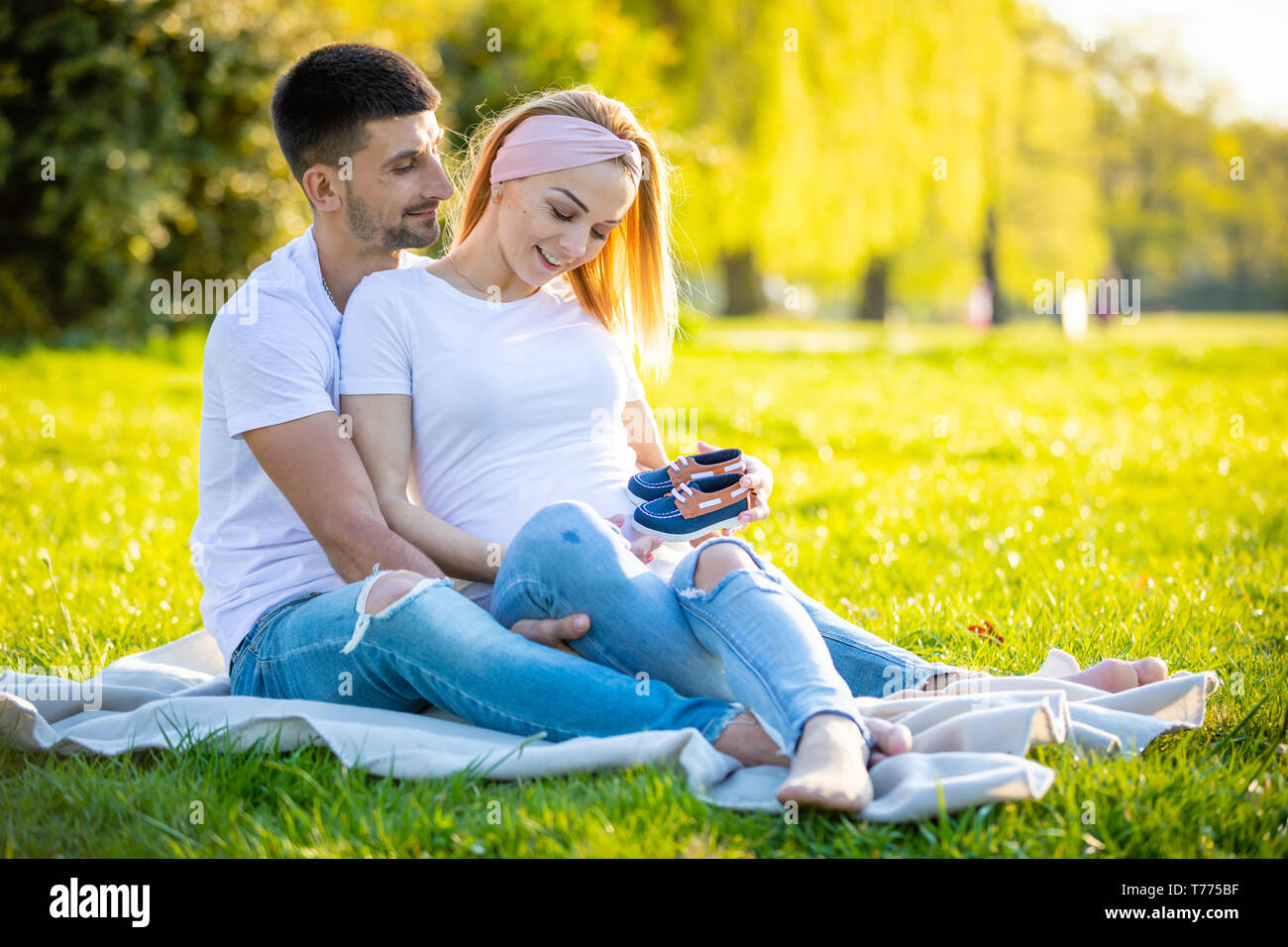 Coppia felice aspetta baby, incinta giovane donna con il marito seduto sul prato verde e azienda Stivali bambino, giovane famiglia e vita nuova nozione Foto Stock