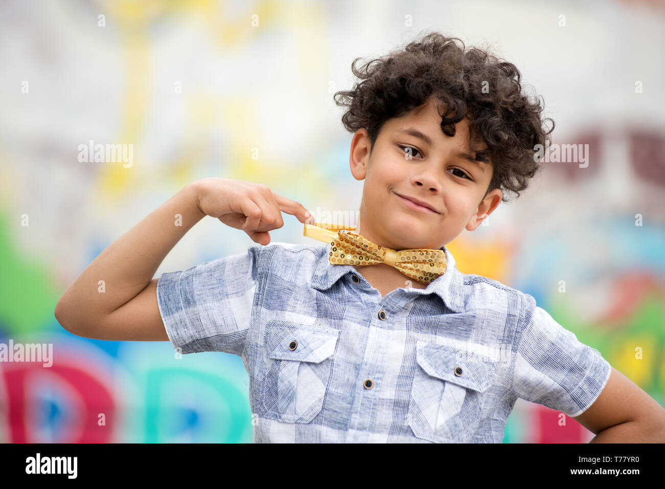 Orgoglioso ragazzo puntando al suo colore oro bow tie con un cordiale sorriso raggiante davanti ad un coloratissimo graffito parete coperta Foto Stock
