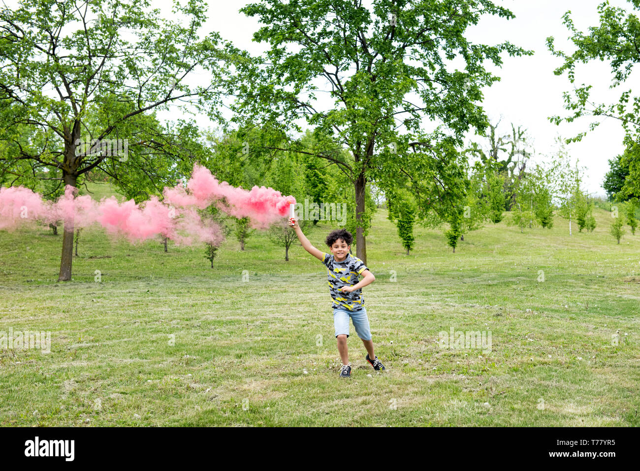 Giovane ragazzo in esecuzione attraverso un parco in primavera si trascina un fumo rosa flare dietro di lui Foto Stock