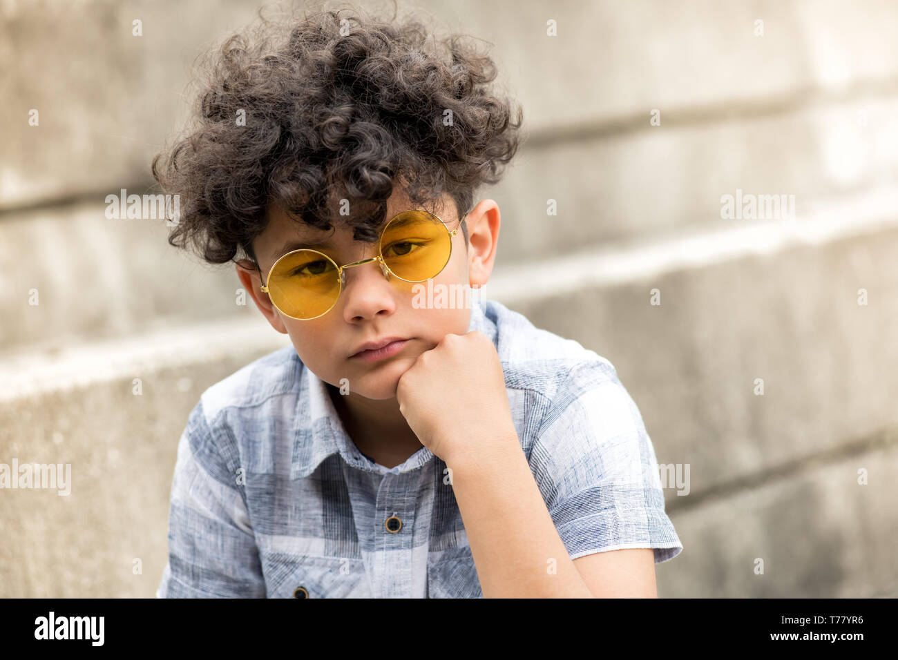 Grave giovane ragazzo con tousled capelli ricci seduto su un gradino all'aperto nel quartiere alla moda di Giallo rotondo occhiali da sole con il mento sulla mano fissando la telecamera Foto Stock