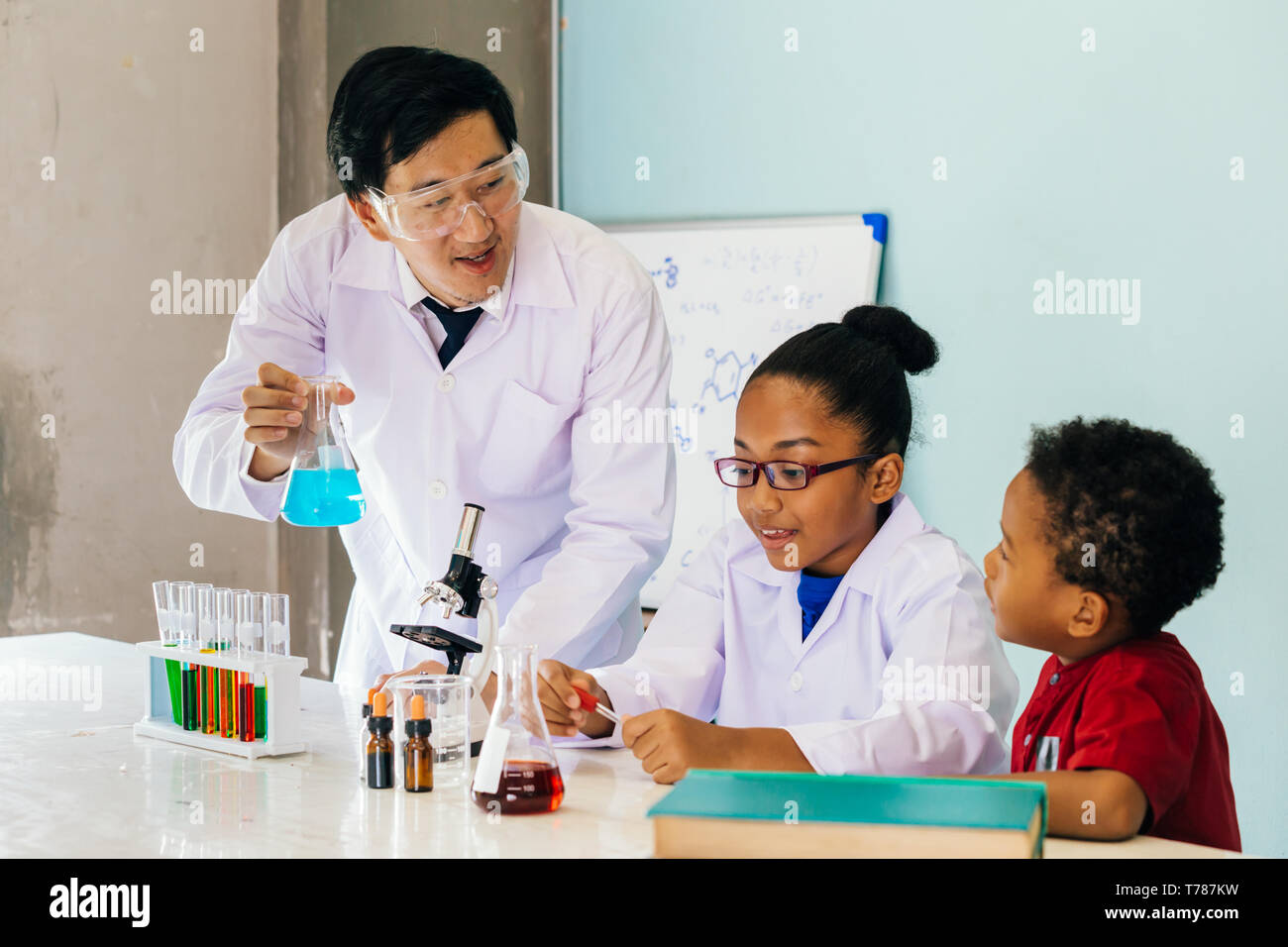 Giovane scienziato asiatico tenendo il tubo di vetro pallone e insegnamento due americano africano bambini misti in laboratorio di chimica esperimento in classe di scienze Foto Stock
