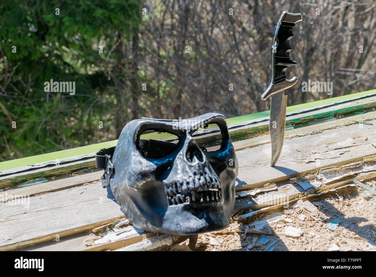 Una maschera terribile nella forma di un cranio e un vecchio coltello da combattimento bloccato nel distrutto davanzale della finestra di una casa abbandonata contro il verde Foto Stock
