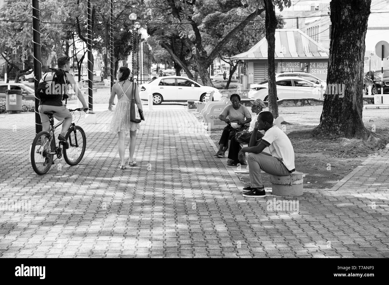 Porto di Spagna, Trinidad e Tobago - Novembre 28, 2015: americano africano gente seduta sul banco in posizione di parcheggio con il giovane coppia di bella ragazza camminare vicino a uomo sportiva in bicicletta per esterno Foto Stock