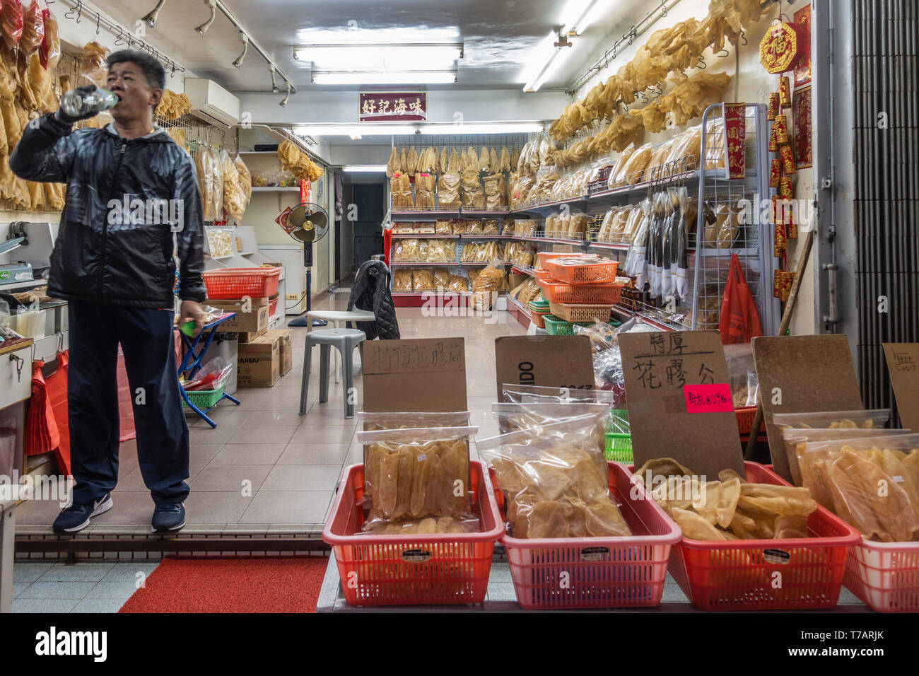 Hong Kong, Cina - 7 Marzo 2019: Tai O villaggio di pescatori. Proprietario di un negozio di vestiti di nero beve l'acqua proteggendo allo stesso tempo il suo grande pesce essiccato store, del display Foto Stock