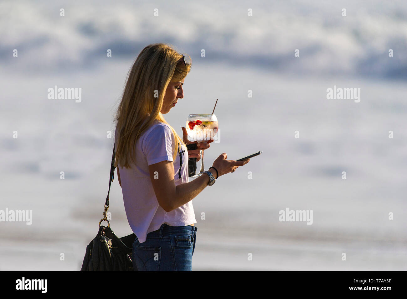 Una donna in piedi di sera la luce del sole tenendo un cocktail e i messaggi di testo sul suo telefono cellulare. Foto Stock