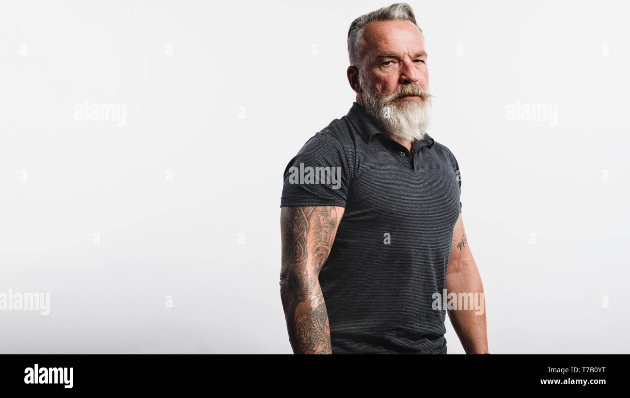 Il vecchio uomo muscoloso con un breve barba bianca guardando la fotocamera. Ritratto di un uomo anziano con tatuaggio su bracci isolati su sfondo bianco. Foto Stock