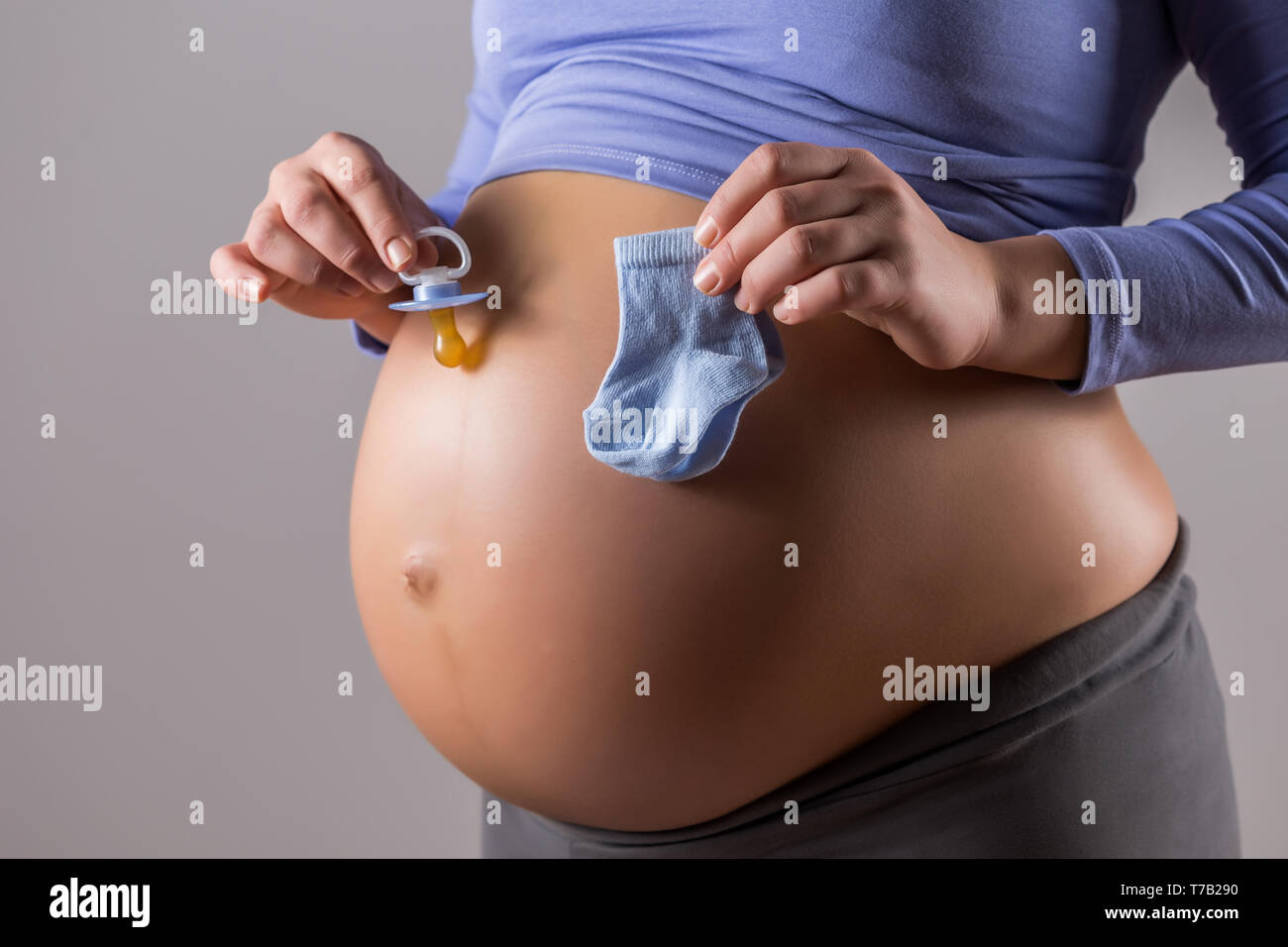 Immagine del ventre della donna incinta con tenendo il succhietto e calze per bambino su sfondo grigio. Foto Stock
