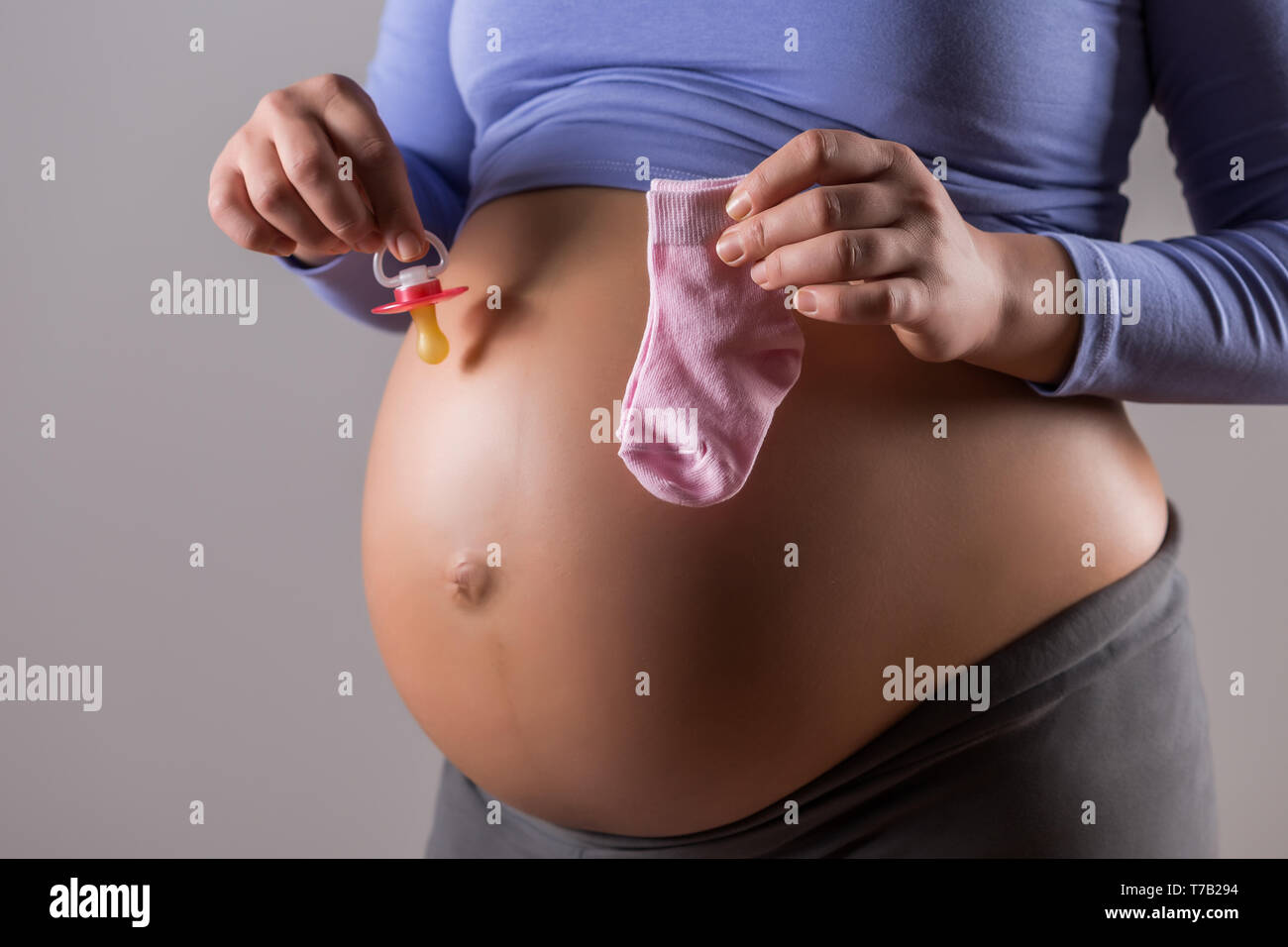 Immagine del ventre della donna incinta con tenendo il succhietto e calzini per bambina su sfondo grigio. Foto Stock