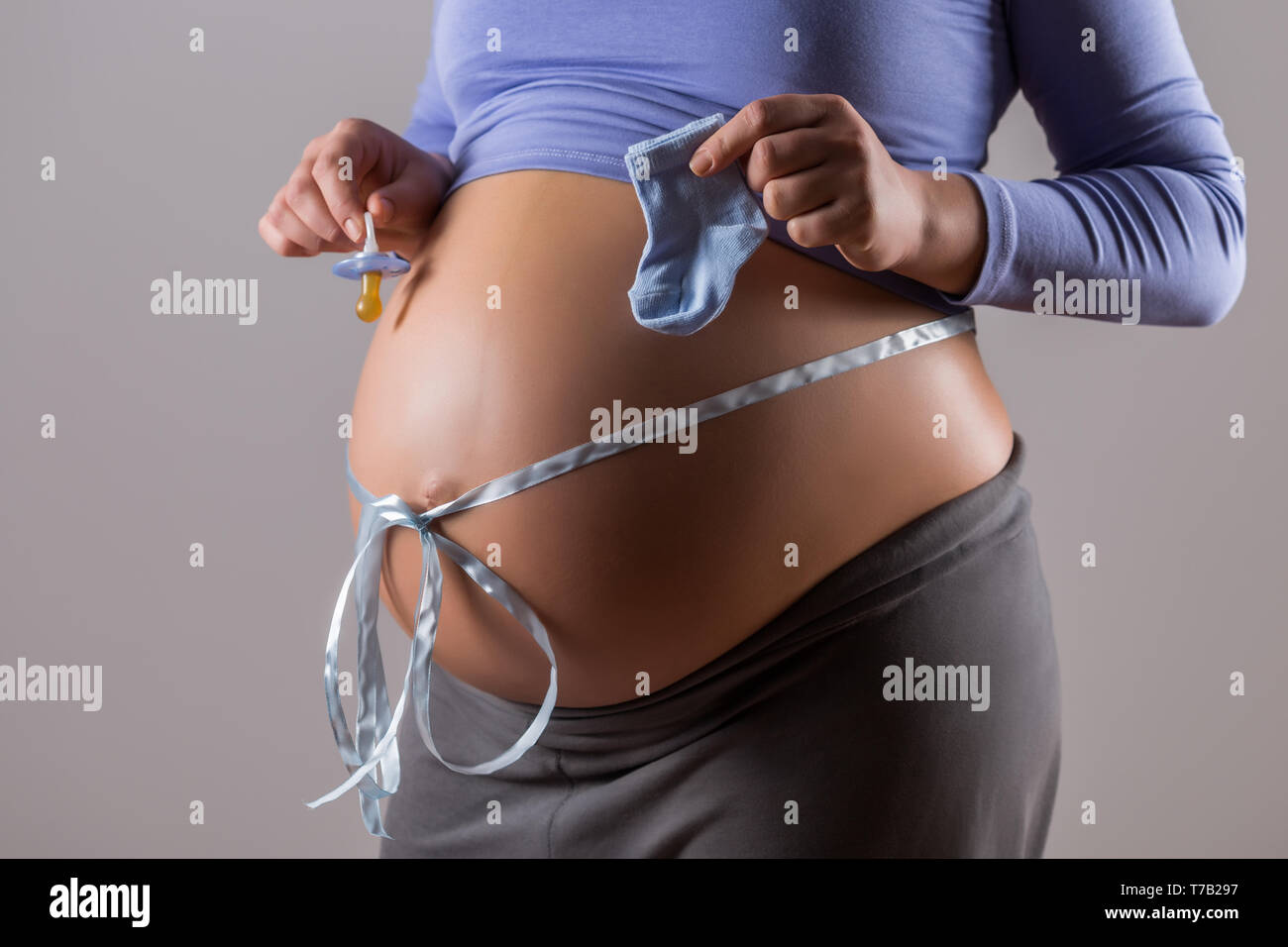 Immagine del ventre della donna incinta con un nastro blu succhietto holding e le calze per bambino su sfondo grigio. Foto Stock