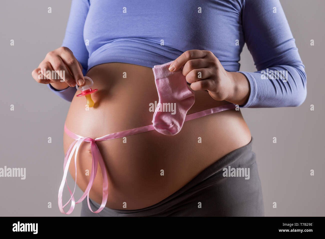 Immagine del ventre della donna incinta con il nastro rosa azienda succhietto e calzini per bambina su sfondo grigio. Foto Stock