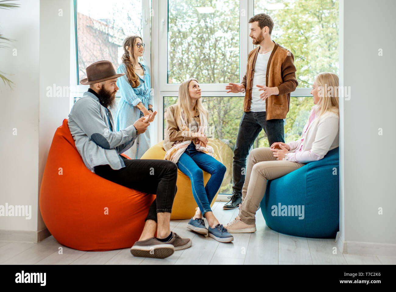 Gruppo di diverse persone aventi una conversazione durante la pausa caffè seduti sul pouf colorato vicino alla finestra in ufficio Foto Stock