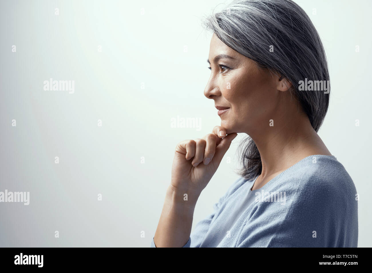 Asian Middle-Aged Profilo di donna. Bella Donna con i capelli grigi si erge su uno sfondo bianco in Studio leggermente sorridente e tocca il suo mento. Ella è T Foto Stock
