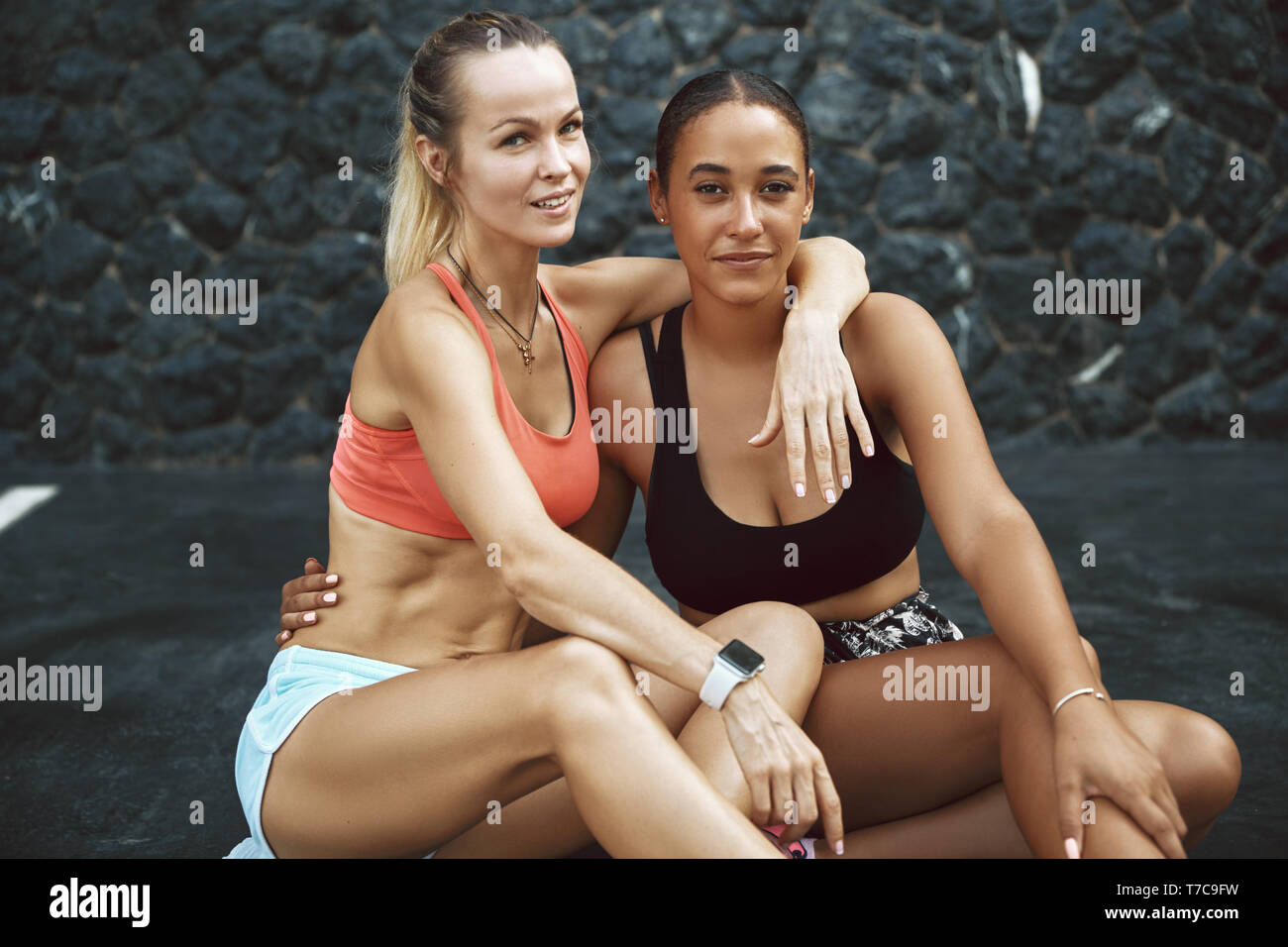 Montare due giovani donne in sportswear sorridere mentre seduto a braccetto in un parcheggio esterno dopo una corsa insieme Foto Stock