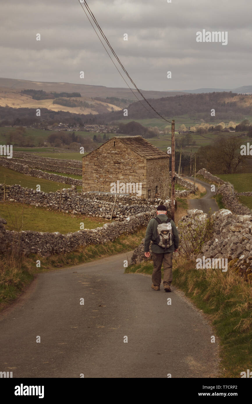 Senior uomo a camminare da soli verso il basso uno stretto vicolo del paese con uno zaino e un vecchio fienile, Wharfedale, Yorkshire Dales, REGNO UNITO Foto Stock