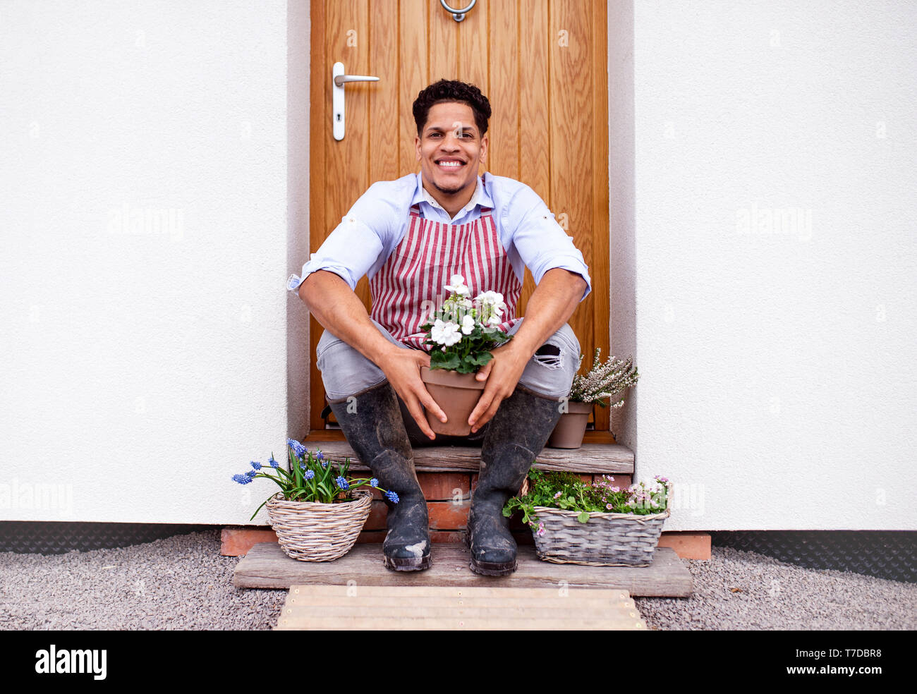 Un Ritratto di giovane allegro uomo seduto davanti alla porta di casa, piantare fiori. Foto Stock
