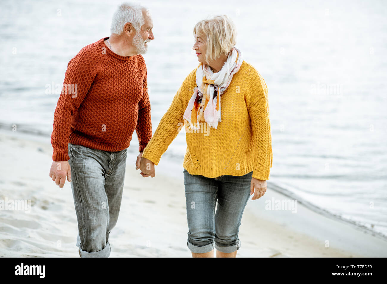 Bella coppia senior vestito in colorate maglioni camminando sulla spiaggia sabbiosa, godersi il tempo libero durante il pensionamento vicino al mare Foto Stock