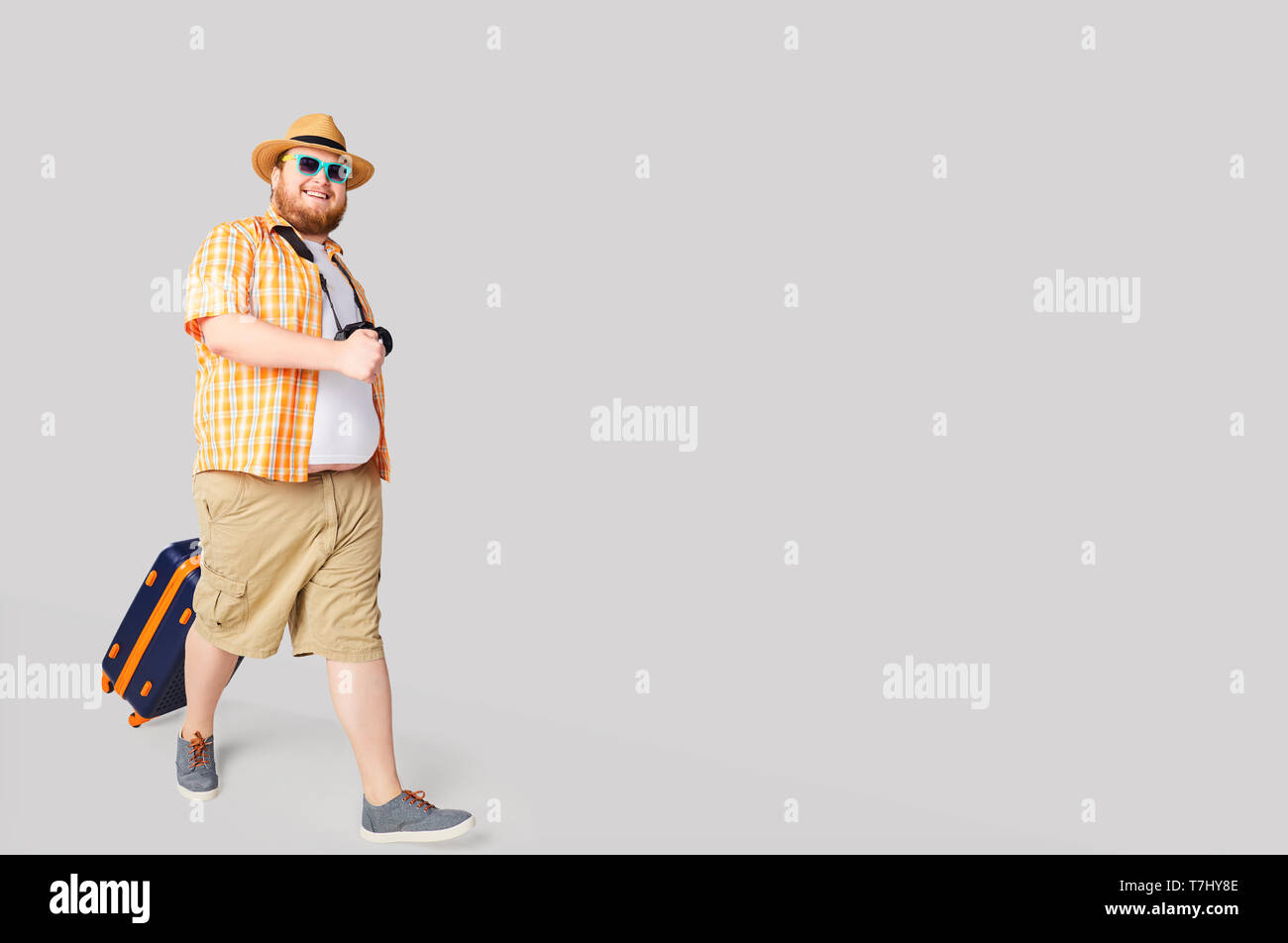 Fat funny man con una valigia sorridente su sfondo grigio. Foto Stock