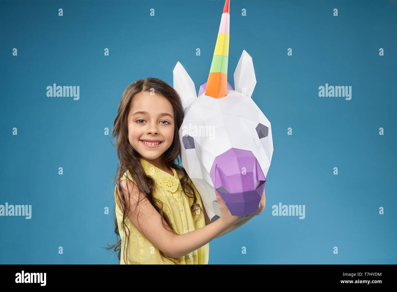 Portret del grazioso e piccolo, modello di felice con la carta unicorn. Bella ragazza positiva in posa,guardando la telecamera, sorridente e tenendo la mano su bianco 3D unicorn testa. Papercraft concetto. Foto Stock
