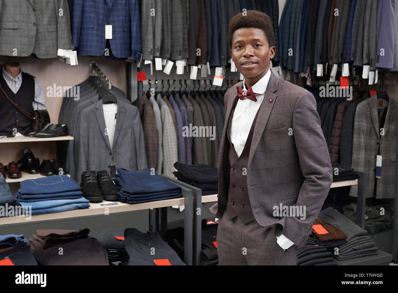 Client di boutique tenendo la mano in tasca dei pantaloni, guardando la fotocamera. Uomo bello il montaggio e posa in abito grigio, camicia bianca e rossa il filtro bow tie. Ampia scelta di abbigliamento moderno su appendiabiti e ripiani. Foto Stock