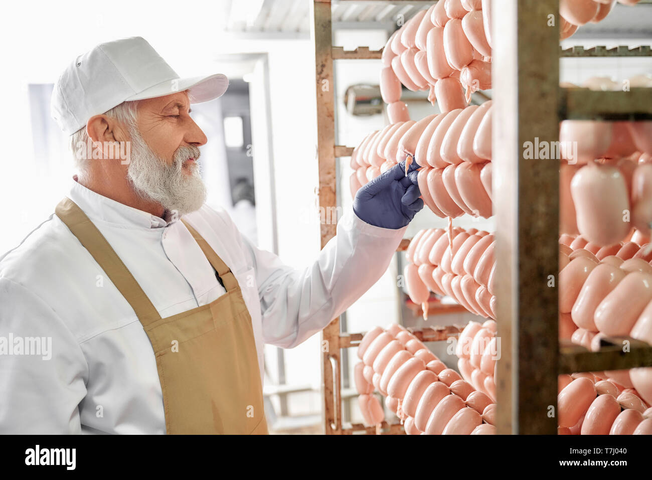 Salsicce di carne di produzione di fabbrica lavoratore. Salsicce appesi in fila. Lavoratore anziano osservazione e controllo, prova salsicce. Uomo in bianco uniforme, tappo bianco, marrone grembiule e guanti di gomma. Foto Stock