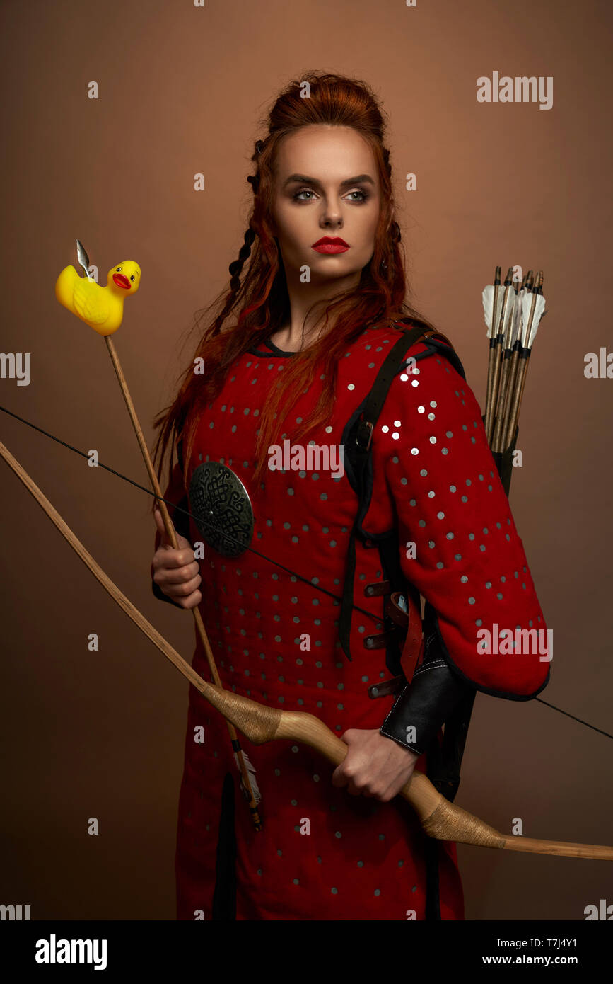 Grave, donna coraggiosa, guerriero medievale tenendo la prua e la freccia con rubber duck. Splendida, bella donna che indossa in tunica rossa che guarda lontano, ponendo in studio. Foto Stock