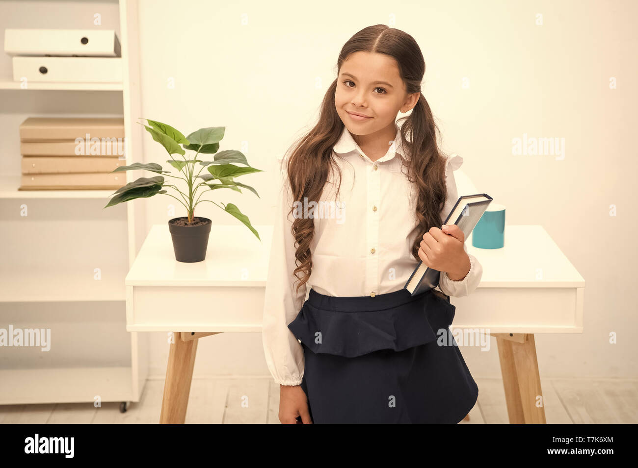 Eccellente prima ex. Bambine tenere libro mentre la tavola di supporto interno bianco. Schoolgirl studiando un libro di testo. Kid uniformi scolastiche faccia felice attesa libro. Entusiasti di conoscenza. Pronto immettere prima forma. Foto Stock