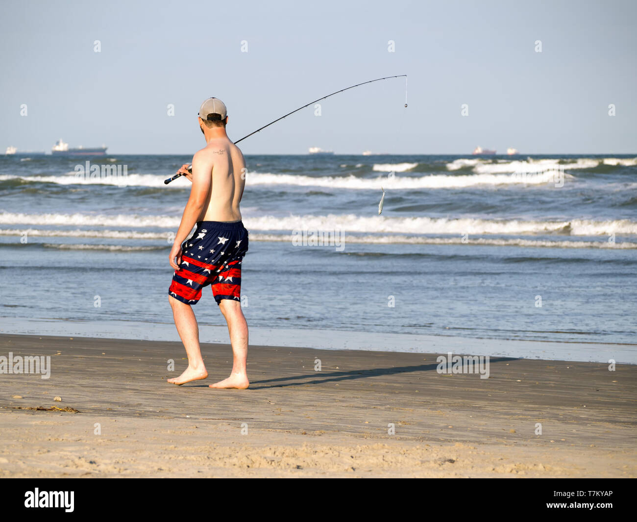 Un pescatore con esche penzolanti dalla pole in pantaloncini corti e tappo a sfera passeggiate sulla spiaggia accanto all'acqua in Port Aransas, Texas, Stati Uniti d'America. Foto Stock