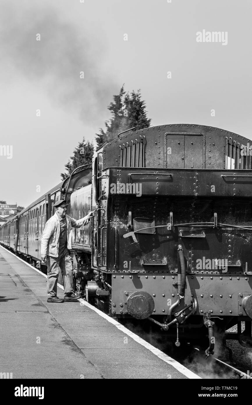 Pilota di equipaggio di treno a vapore bianco e nero, in berretto e tuta in fiocchi, su piattaforma ferroviaria che si imbarcarsi in attesa di una locomotiva a vapore britannica d'epoca, estate. Foto Stock