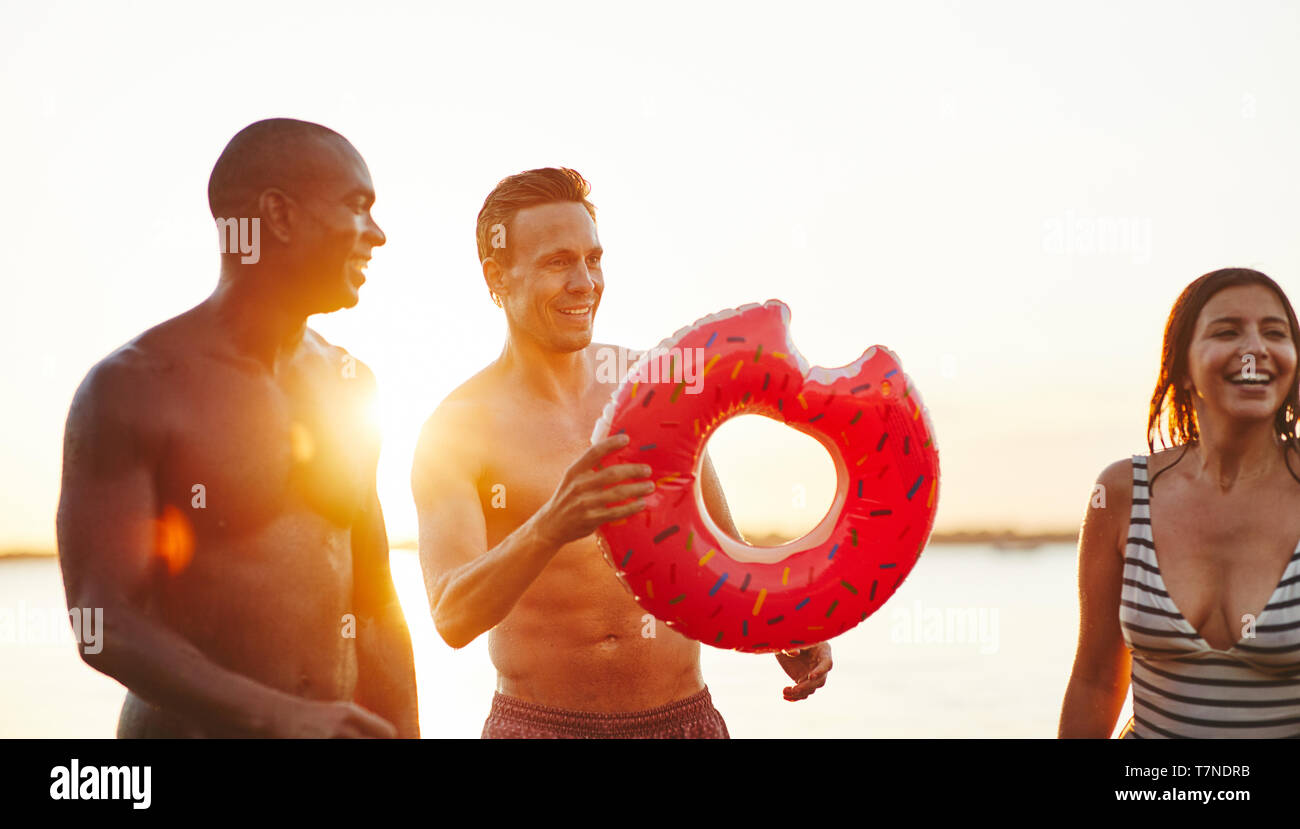Sorridente giovane in un costume da bagno in possesso di un anello di nuotare divertendosi con un gruppo di diversi amici presso un lago Foto Stock