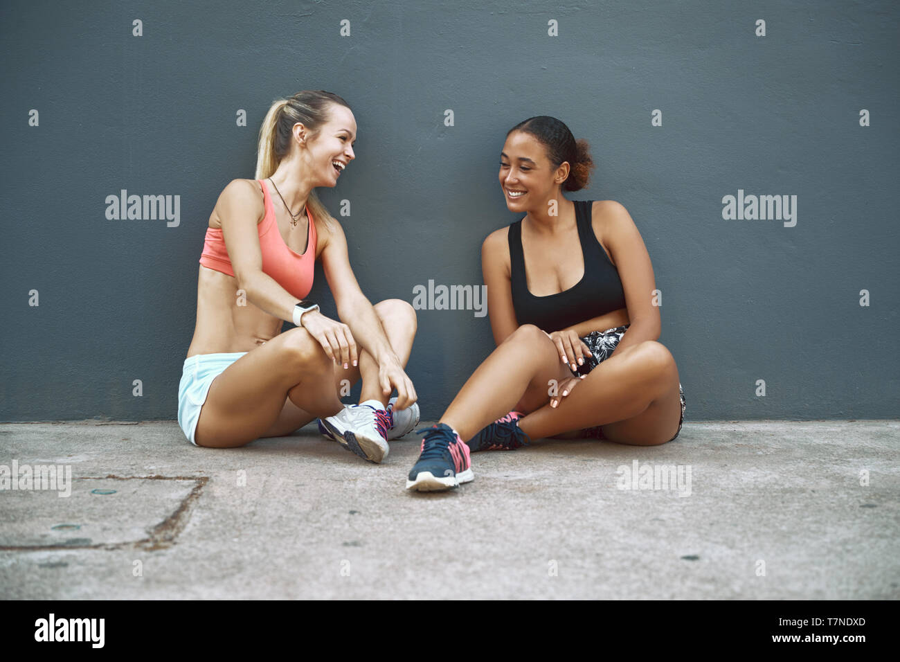 Montare due giovani donne in sportswear seduta sul terreno al di fuori di parlare e ridere mentre prendendo una pausa dal loro allenamento Foto Stock