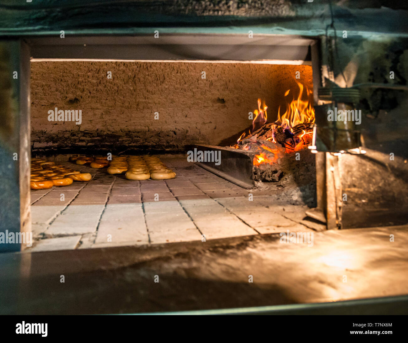 Fuoco e brace ardente riscaldano il forno a legna in mattoni di una panetteria tradizionale a Dobrich, in Bulgaria. Diversi filetti di anelli di gevrek dorati e marroni stanno già cuocendo direttamente sul fondo del forno, senza alcun vassoio. L'officina probabilmente appartiene al museo all'aperto Stariyat Dobrich (Vecchia Dobrich), che ha ricreato l'architettura storica del mercato della città dalla fine degli anni '70/inizio degli anni '80 e riunisce più di 30 laboratori artigianali, tra cui questa panetteria di gevrek con il suo forno a legna. Foto Stock