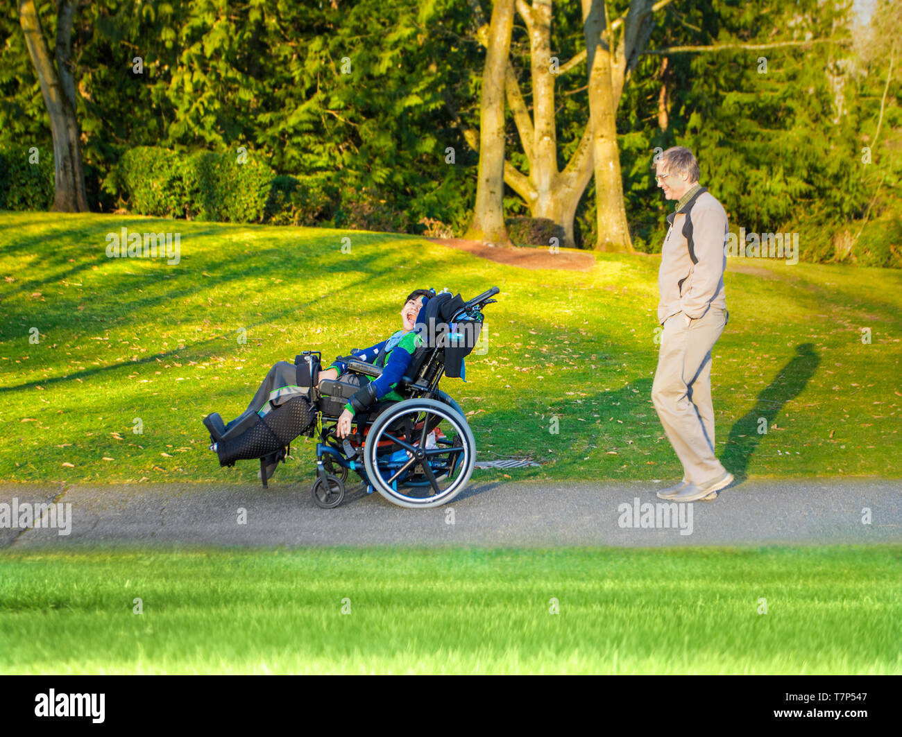 Padre caucasica camminare dietro disabilitato biracial giovane figlio al parco all'aperto in primavera o in estate. Bambino ha paralisi cerebrale. Foto Stock